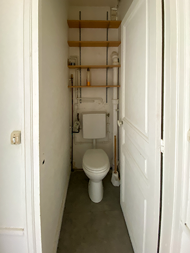 Salle de bain avant les travaux avec une latrine posée de travers avec un réservoir et des tuyaux apparents.