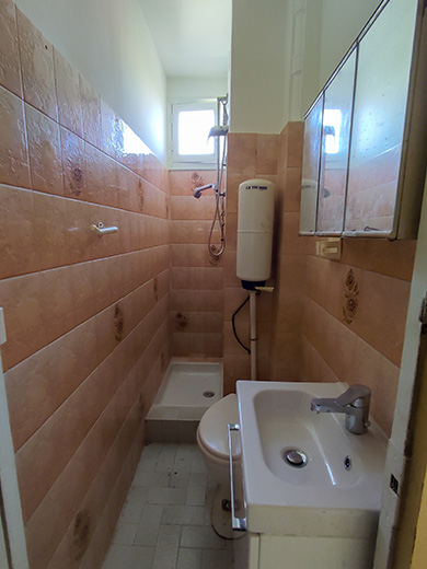 Salle de bain à rénover avec un carrelage en dégradé de rose des années 1970, un toilette avec son réservoir apparent dans l'angle de la pièce, et une douche avec un rideau.