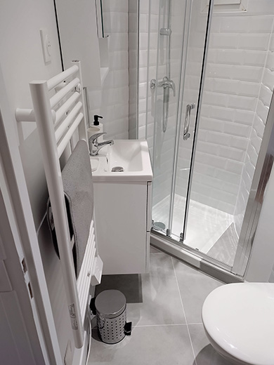 Salle de bain moderne et lumineuse avec des carreau de métro biseautés blancs, une cabine de douche avec porte coulissante, un petit lavabo d'appoint et un WC suspendu.