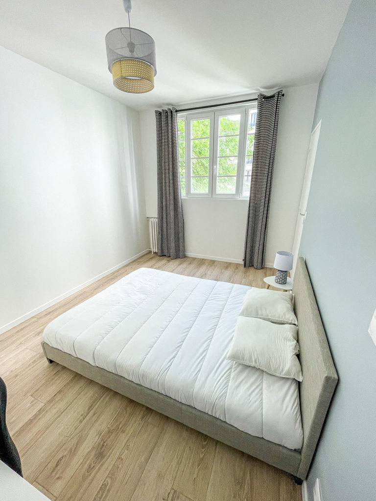 Chambre après les travaux avec un mur contrastant bleu pastel, le reste des murs en blanc, un parquet en bois clair, un lit gris et des rideaux noirs avec un motif à pied de poule gallois